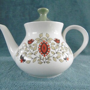 Puede incluir: Tetera blanca con tapa verde y un patrón floral en tonos rojos, marrones y beige.