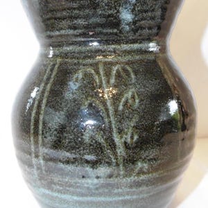 Puede incluir: Un jarrón de cerámica hecho a mano con un esmalte oscuro y moteado en tonos verdes y negros. El jarrón presenta un cuerpo bulboso y un borde acampanado. Patrones florales decorativos están grabados en la superficie.