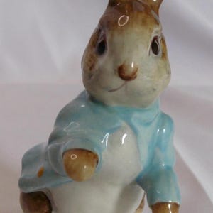 May include: A ceramic rabbit figurine wearing a light blue jacket and white trousers, standing on a green base. The rabbit has brown ears and a light brown nose. A collectible item.