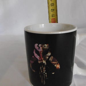 May include: A black ceramic mug with a white interior and handle. The mug features a colorful graphic of a character from a science fiction franchise. The mug is approximately 4 inches tall.