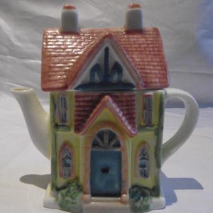 Peut inclure: Théière en céramique en forme de maison. Le toit est rouge avec un motif de briques, et la maison est peinte en jaune avec une porte et des fenêtres bleues. Elle a une anse et un bec blancs, et deux cheminées.