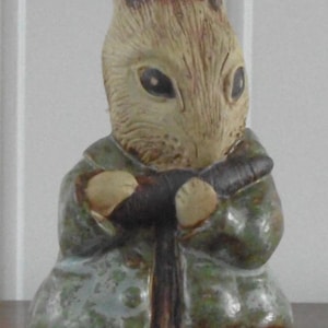 May include: A ceramic figurine of a rabbit wearing a green coat. The rabbit has brown and tan fur, with its arms crossed. The coat is a muted green with a slightly weathered appearance. The figurine is on a wooden surface.