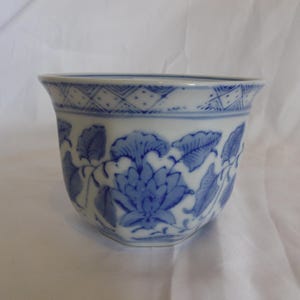 May include: A white porcelain bowl featuring a blue underglaze floral pattern with detailed leaves and petals. The rim is decorated with a geometric cross-hatch border. The bowl has a flared opening and a hexagonal base, showcasing a classic design.