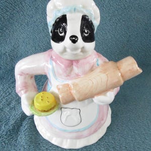 May include: A ceramic teapot shaped like a badger wearing a pink and white apron and a white chef's hat. The badger is holding a rolling pin and a plate with a yellow pastry on it.