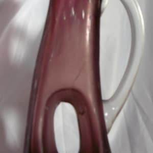 May include: A tall, purple glass jug with a white handle. The jug has a unique, elongated shape with a large oval opening in the centre. The glass has small white bubbles.