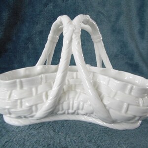 Puede incluir: Cesta de cerámica blanca con un diseño tejido y dos asas. La cesta tiene forma de semicírculo y un acabado liso y brillante.