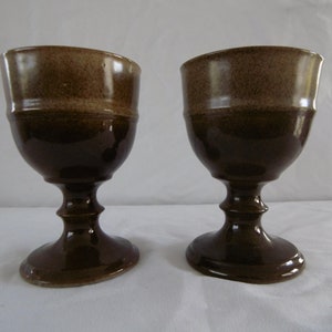 May include: Two brown ceramic goblets with a light brown band around the top of the bowl. The goblets have a rounded bowl and a tall, slender stem.