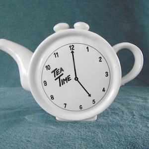 Puede incluir: Tetera de cerámica blanca con forma de reloj con agujas negras y la inscripción "Tea Time" en la esfera.