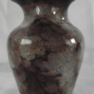 May include: A ceramic vase with a speckled, mottled finish in shades of brown, grey, and burgundy. The vase has a flared rim and a rounded body, suitable for displaying flowers or as a decorative piece.