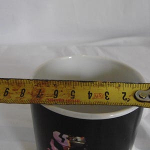 May include: A black and white ceramic mug with a white interior. A yellow tape measure is extended across the top of the mug, showing a measurement of approximately 3 inches.