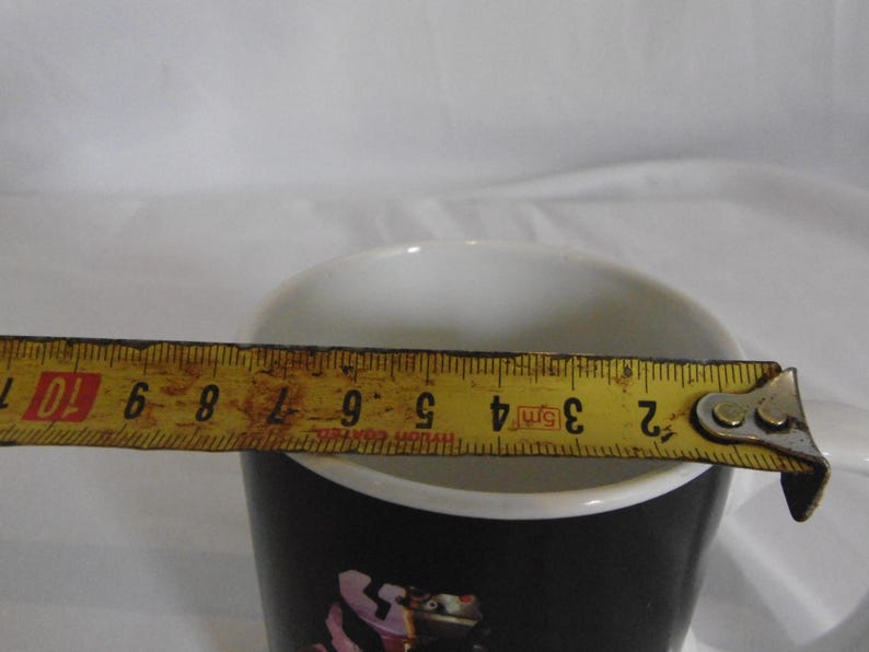 May include: A black and white ceramic mug with a white interior. A yellow tape measure is extended across the top of the mug, showing a measurement of approximately 3 inches.