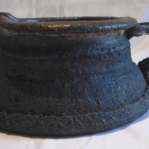 May include: A dark, textured ceramic pitcher with a handle and a spout. The pitcher has a wide base and a narrow top, with a rough, uneven surface. The handle is curved, and the spout is slightly angled. The overall color is a dark charcoal.