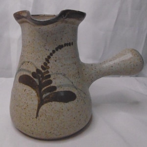 May include: A speckled ceramic jug with a brown handle and a decorative brown leaf design. The jug has a ruffled rim and a light beige colour. The jug is a unique piece of pottery.