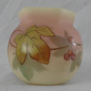 May include: A small, round, pale yellow vase with a pink upper section. The vase is decorated with a large yellow and brown leaf and a cluster of red berries. Green leaves and stems are also painted on the vase.