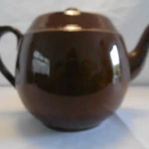 Könnte beinhalten: Eine braune Teekanne aus Keramik mit rundem Korpus und einem einfachen Henkel. Die Teekanne hat eine glänzende Oberfläche.