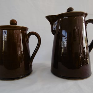May include: Two brown ceramic coffee pots with lids and handles. The larger pot is taller than the smaller pot.
