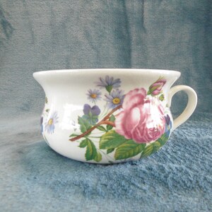 Op de afbeelding: Een witte keramische chamberpot met een bloemenontwerp met roze rozen, blauwe en paarse bloemen en groene bladeren.