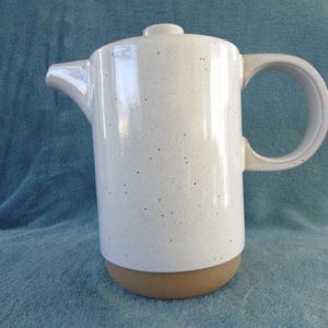 Könnte beinhalten: Weiße Kaffeekanne aus Keramik mit gesprenkeltem Glase und braunem Sockel. Die Kanne hat einen Henkel und einen Ausguss.
