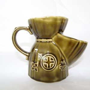 Puede incluir: Una taza de afeitar de cerámica verde con asa y pico. La taza tiene un diseño decorativo con una cruz y flores.