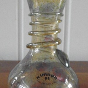 Puede incluir: Un jarrón decorativo de vidrio con base bulbosa y parte superior acampanada. El jarrón presenta un diseño en espiral alrededor del cuello y un acabado metálico con tonos amarillos, marrones y rojos. El texto "Nimicon" es visible.