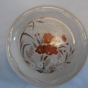 Peut inclure: Une assiette à tarte en céramique blanche avec un bord festonné et un motif floral composé de fleurs rouges et brunes et de feuilles blanches et brunes.