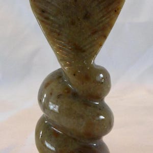 May include: A carved stone cobra statue. The sculpture features a detailed cobra head with an open hood, atop a coiled body. The stone has a mottled, earthy color with variations of brown and tan.