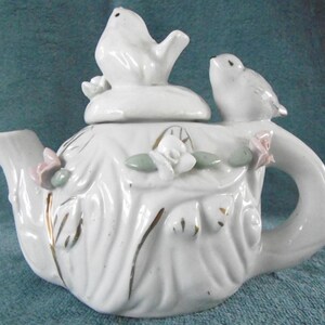 Puede incluir: Tetera de porcelana blanca con un pájaro posado en la tapa y otro pájaro en el asa. La tetera está decorada con detalles dorados y flores rosas y verdes.