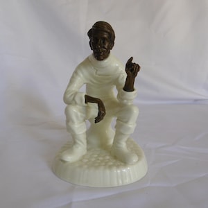 May include: A figurine of a person in a seated position, featuring a dark brown head and hands contrasted against a white outfit and base. The figure wears a high-collared top and boots, with one hand raised in a gesture, set upon a textured circular base.