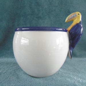 May include: White ceramic bowl with a blue rim and a toucan handle. The toucan is painted with yellow, blue, and black details. The bowl has a glossy finish and a round shape.