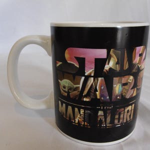 May include: A black ceramic mug with a white handle. The mug features the words "STAR WARS" in a multi-colored design, with "THE MANDALORIAN" below. The mug has a white interior.