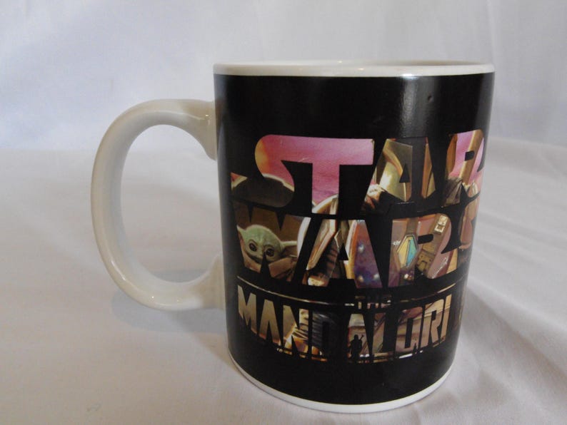May include: A black ceramic mug with a white handle. The mug features the words "STAR WARS" in a multi-colored design, with "THE MANDALORIAN" below. The mug has a white interior.