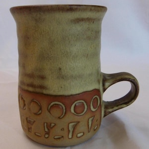 May include: A handmade ceramic mug with a unique design. The mug features a light green upper section and a lower section with a reddish-brown band and carved geometric shapes. The handle is a matching earthy brown color.