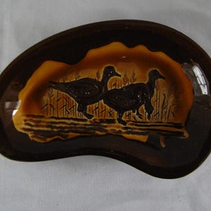 May include: Brown ceramic ashtray with a unique shape. The ashtray features a raised relief design of two ducks in a marsh setting, with brown and tan tones. The rim is a darker brown.
