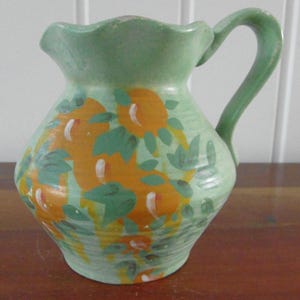 Op de afbeelding: Handbeschilderde keramische kan in lichtgroen met een oranje bloemenontwerp en groene bladeren. De kan heeft een geschulpte rand en een gebogen handvat. De decoratie met oranje bloemen en witte accenten.