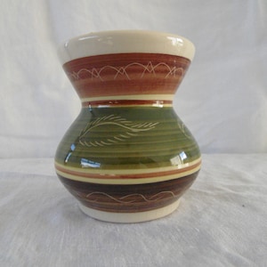 Puede incluir: Un pequeño jarrón de cerámica verde y marrón con un borde blanco y un diseño de hojas estilizadas. El jarrón tiene una boca ancha y una base estrecha.