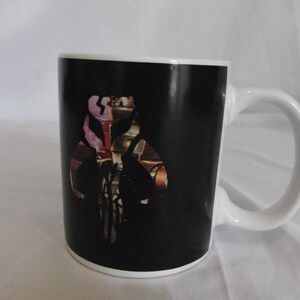 May include: White ceramic coffee mug with a black background featuring a detailed graphic design. The design includes a stylized image with brown, gold, and pink accents. The mug has a white handle and rim.