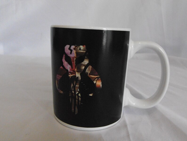 May include: White ceramic coffee mug with a black background featuring a detailed graphic design. The design includes a stylized image with brown, gold, and pink accents. The mug has a white handle and rim.