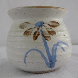 May include: A small, off-white ceramic vase with a hand-painted floral design. The flower has brown petals and a blue centre, with a blue stem and leaves. The vase has a slightly flared rim and a textured surface.