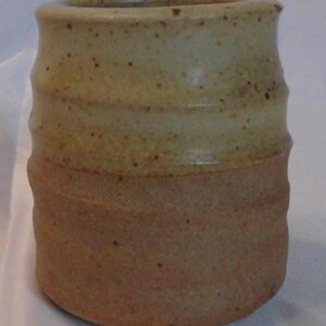 May include: A handmade ceramic tumbler with a speckled, two-tone design. The top half is a light tan colour, while the bottom half is a darker brown. The cylindrical shape has a slightly textured surface.