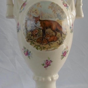 May include: Cream-colored ceramic vase with a fox family scene. The vase has a square base, a bulbous body, and two handles. Floral accents are scattered across the surface. The central image depicts a mother fox with her kits.