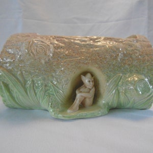 Może przedstawiać: Ceramiczna doniczka w kształcie pnia drzewa z zieloną i brązową glazurą. Mały, biały ceramiczny gnom siedzi w zagłębieniu w pniu.