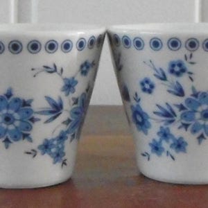 Peut inclure: Quatre verres à shot en céramique blanche avec des motifs floraux bleus. Chaque verre présente un motif floral bleu et une bordure de cercles bleus près du bord. Les verres sont disposés côte à côte sur une surface en bois.
