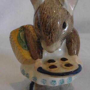 Könnte beinhalten: Keramikfigur einer Maus, die ein Tablett mit Keksen und einen Korb hält. Die Maus ist braun und weiß und trägt eine weiße Schürze mit blauen Blumenmustern. Die Kekse sind braun mit einer roten Füllung.