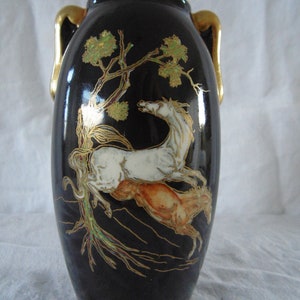 Könnte beinhalten: Eine schwarze Keramikvase mit goldenen Akzenten und einer bemalten Szene von zwei weißen Pferden, die durch einen grünen Wald rennen. Die Vase hat zwei goldene Henkel.