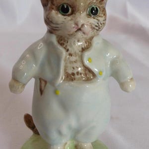 May include: A ceramic cat figurine wearing a light blue outfit with yellow buttons. The cat has brown and tan fur and green eyes, standing on a green base. The figurine is approximately 10 cm tall.