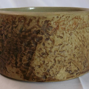 May include: A cylindrical ceramic bowl with a textured, bark-like exterior. The bowl has a light brown and dark brown colour palette, with a smooth, light green interior. The vessel is suitable for decorative or functional use.