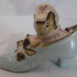 May include: A ceramic figurine of a mouse wearing a hat and scarf, sitting in a light blue shoe. The shoe has a small heel and is decorated with brown accents. The mouse has a detailed face and is surrounded by other mice.