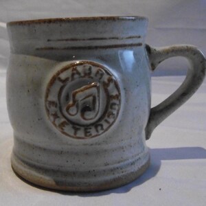 May include: A stoneware mug with a light gray glaze and a brown rim and base. The mug features a circular emblem with a musical note and the text "LAURA ETERIUS MUSIC". The handle is curved and the mug has a rustic, handmade appearance.