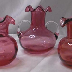 May include: Three cranberry-coloured glass jugs with clear glass handles. Each jug has a ruffled rim and a rounded body. The jugs are arranged in a row, showcasing their design and colour.