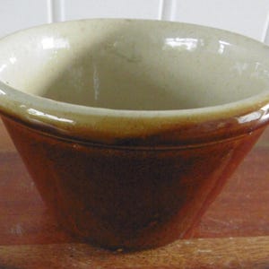 Puede incluir: Un cuenco de cerámica marrón y beige. El cuenco tiene forma redondeada y un acabado brillante. El exterior es de color marrón oscuro, mientras que el interior es de color beige claro. El cuenco está sobre una superficie de madera.